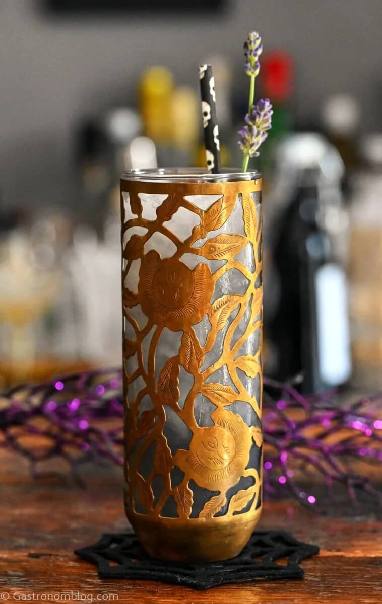 A glass with a gold metal floral decoration on the outside holds a pale purple drink.