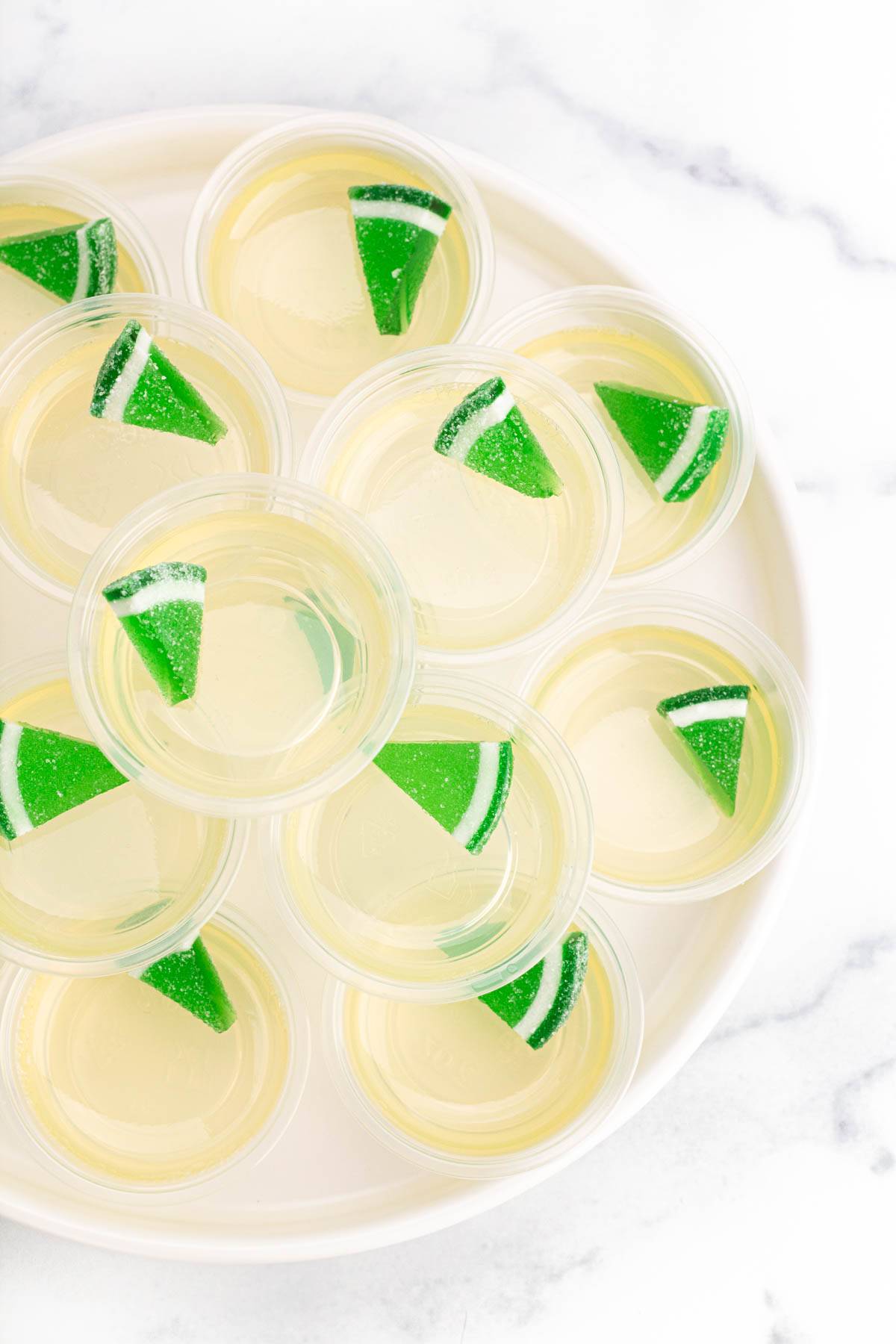 Jello shots sit in clear cups on a round white tray garnished with green candy wedges.