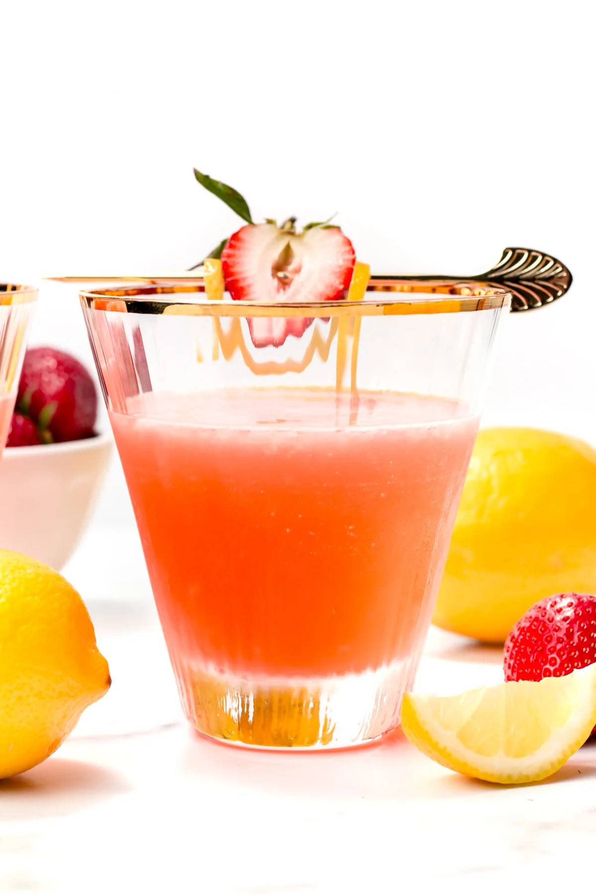 A pink cocktail in a stemless martini glass with a gold rim garnished by half a strawberry and a lemon twist on a gold skewer.