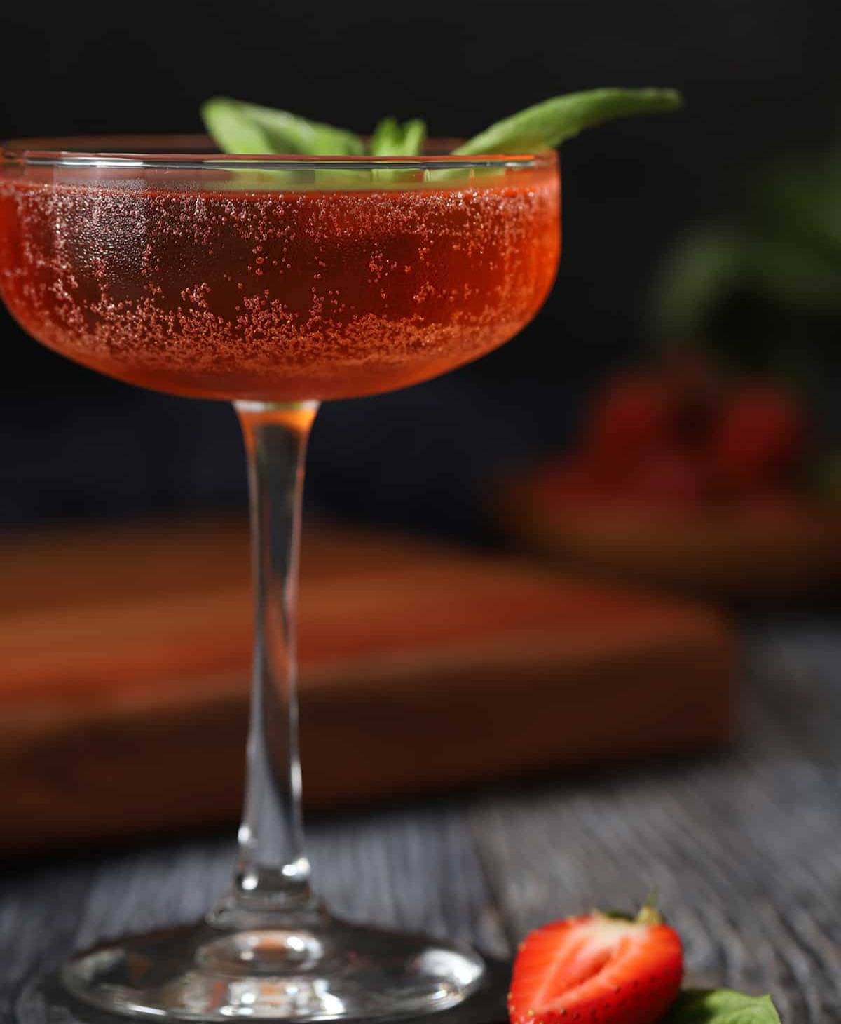 A red cocktail in a coupe glass topped with a sprig of herb.
