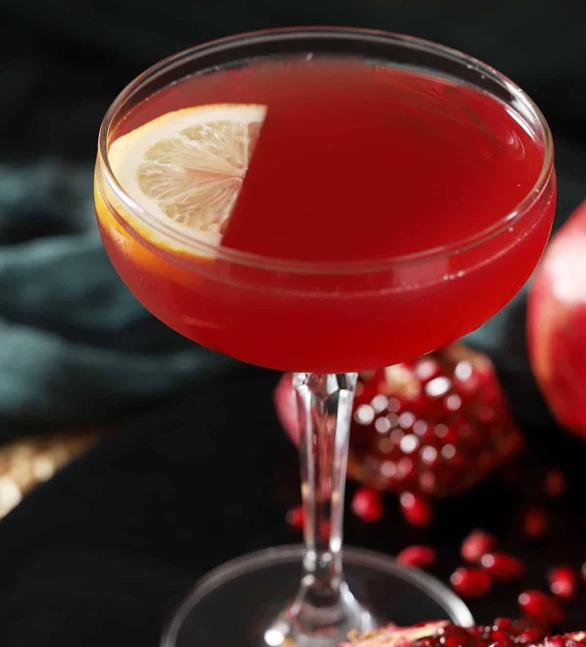 A deep red drink in a stemmed coupe glass with a slice of lemon floating on top.