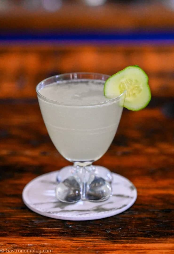 A short glass holds a pale drink with a cucumber round on the rim.