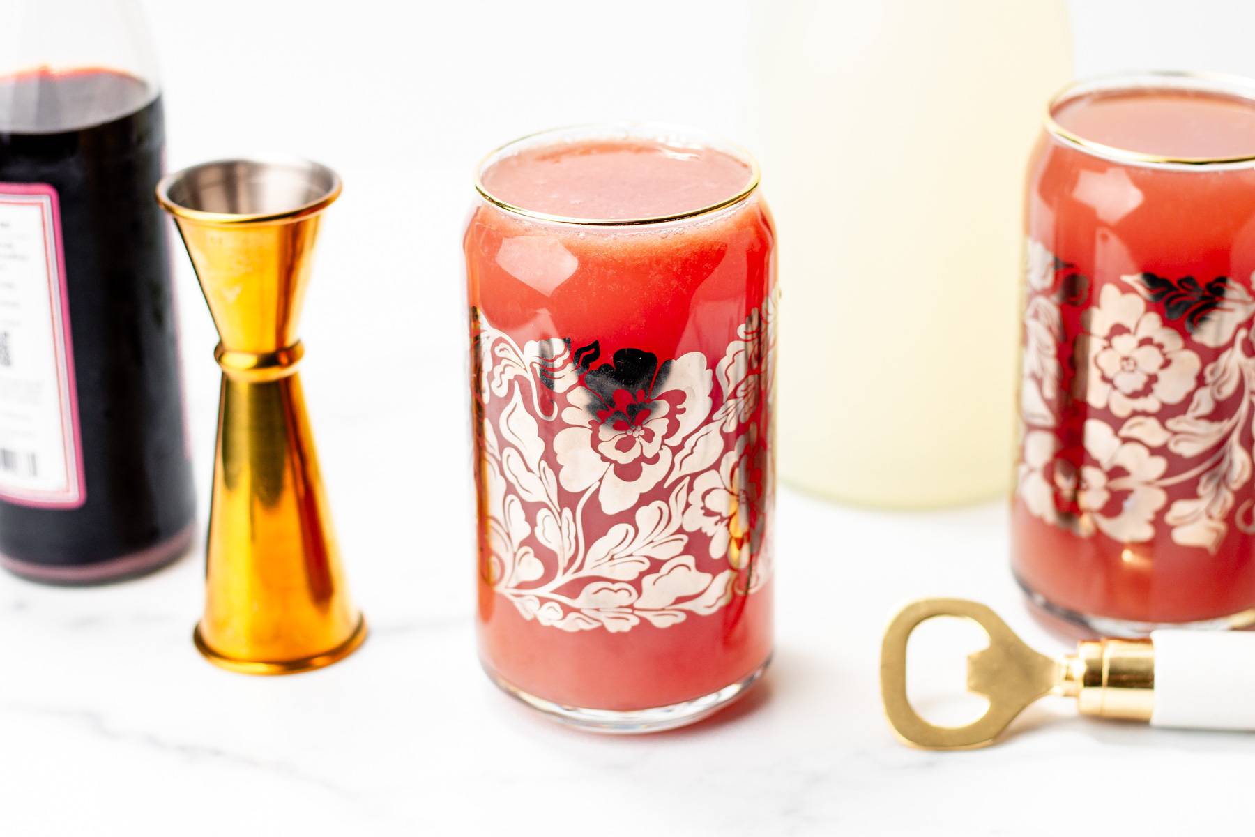 Two can glasses with silver flowers holds a red cocktail near gold bar tools.