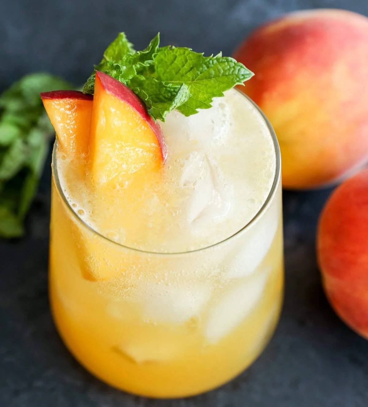A stemless glass holds a light orange drink topped with peach slices and an herb sprig.