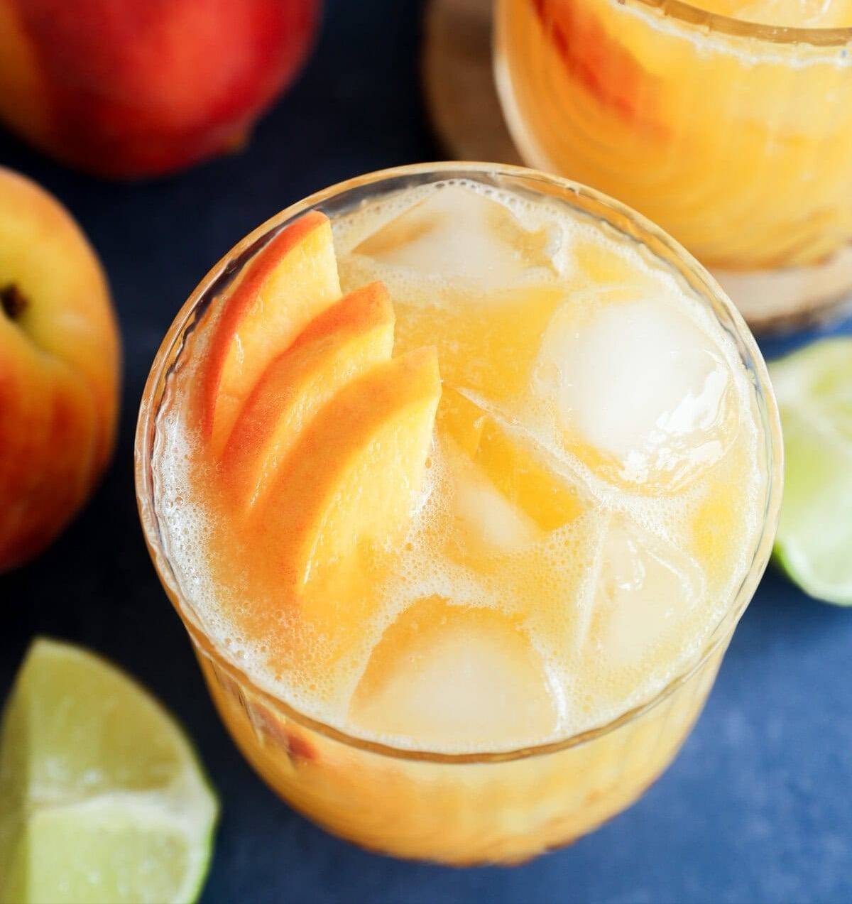 Overhead view of a pale orange cocktail with peach slices and ice cubes near slices of lime and whole peaches.