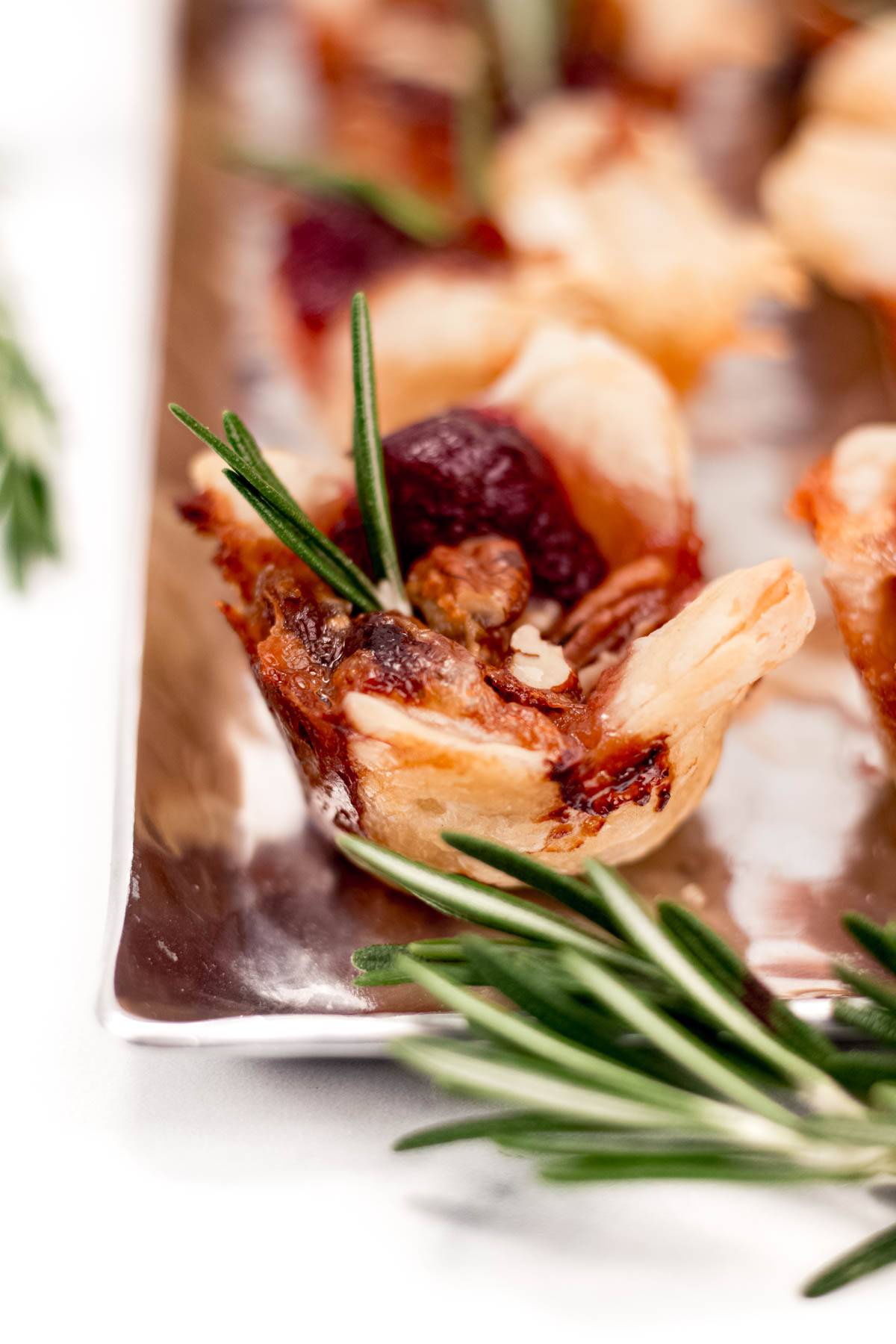 A cranberry brie bite garnished with rosemary on a silver tray.