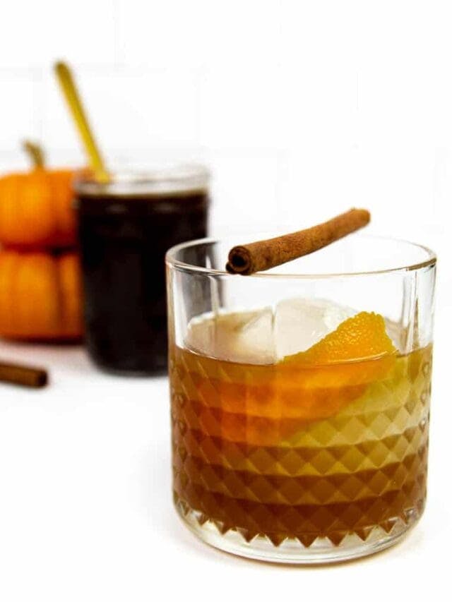 A pumpkin spice old fashioned garnished with a cinnamon stick sits in front of a jar of deep brown pumpkin syrup on a white background.