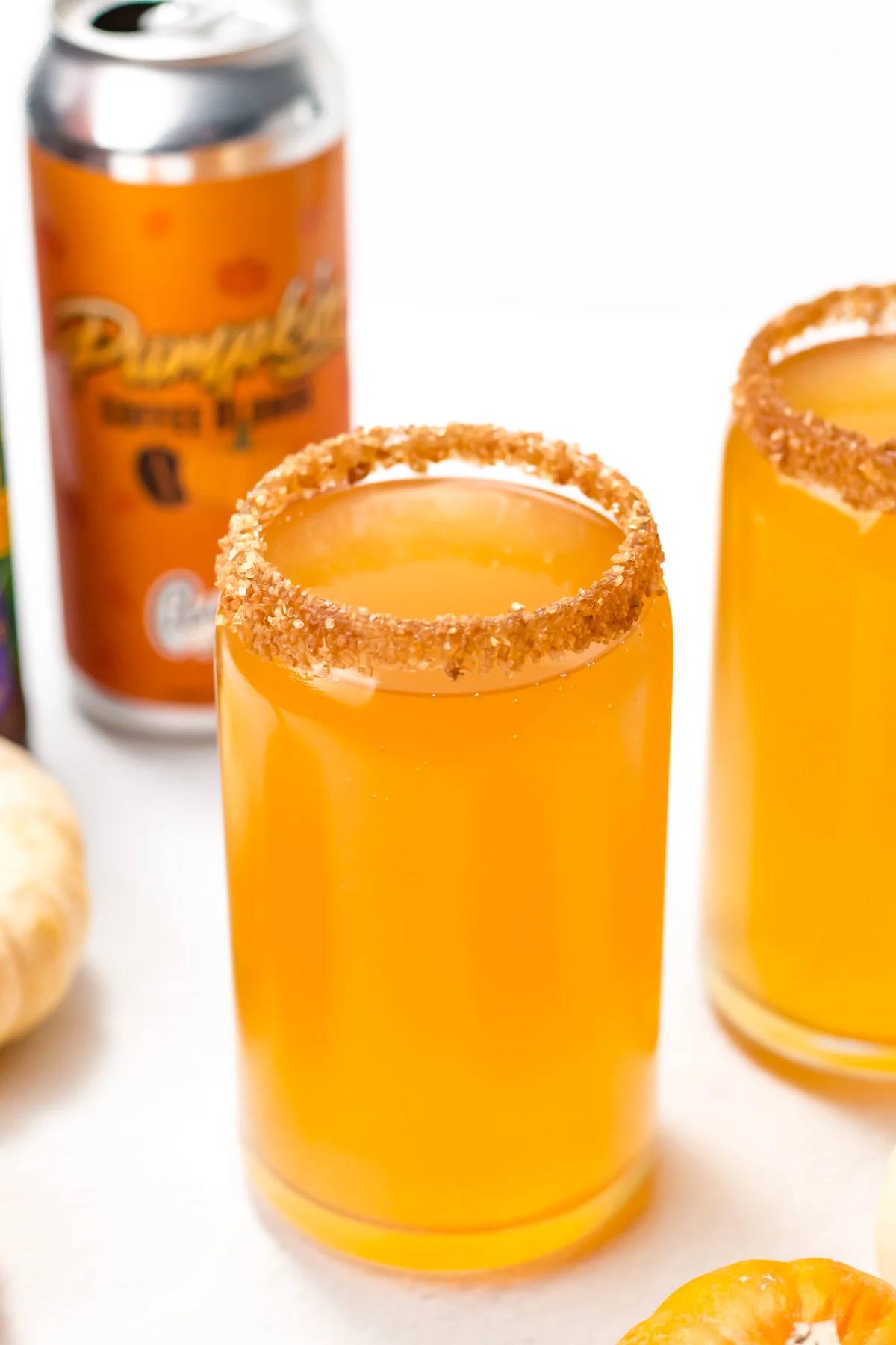 A pale orange drink in a beer can mug rimmed with cinnamon sugar sits in front of another, with a can of pumpkin beer behind.