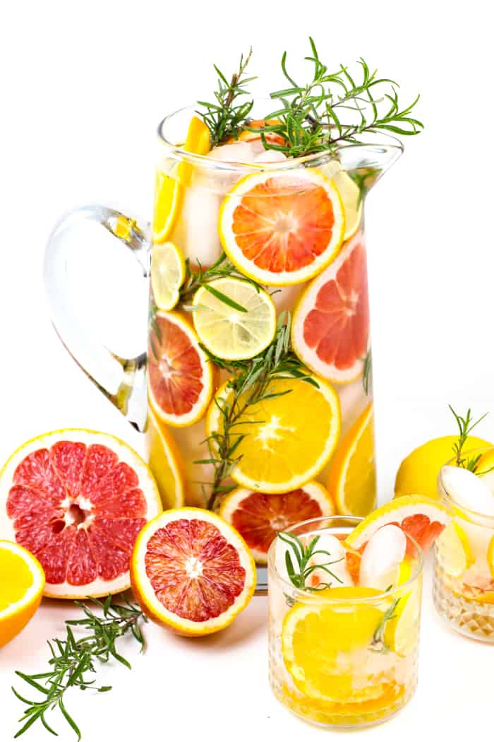 A pitcher filled with citrus and herb sprigs sits near two glasses of water with citrus and herb sprigs.