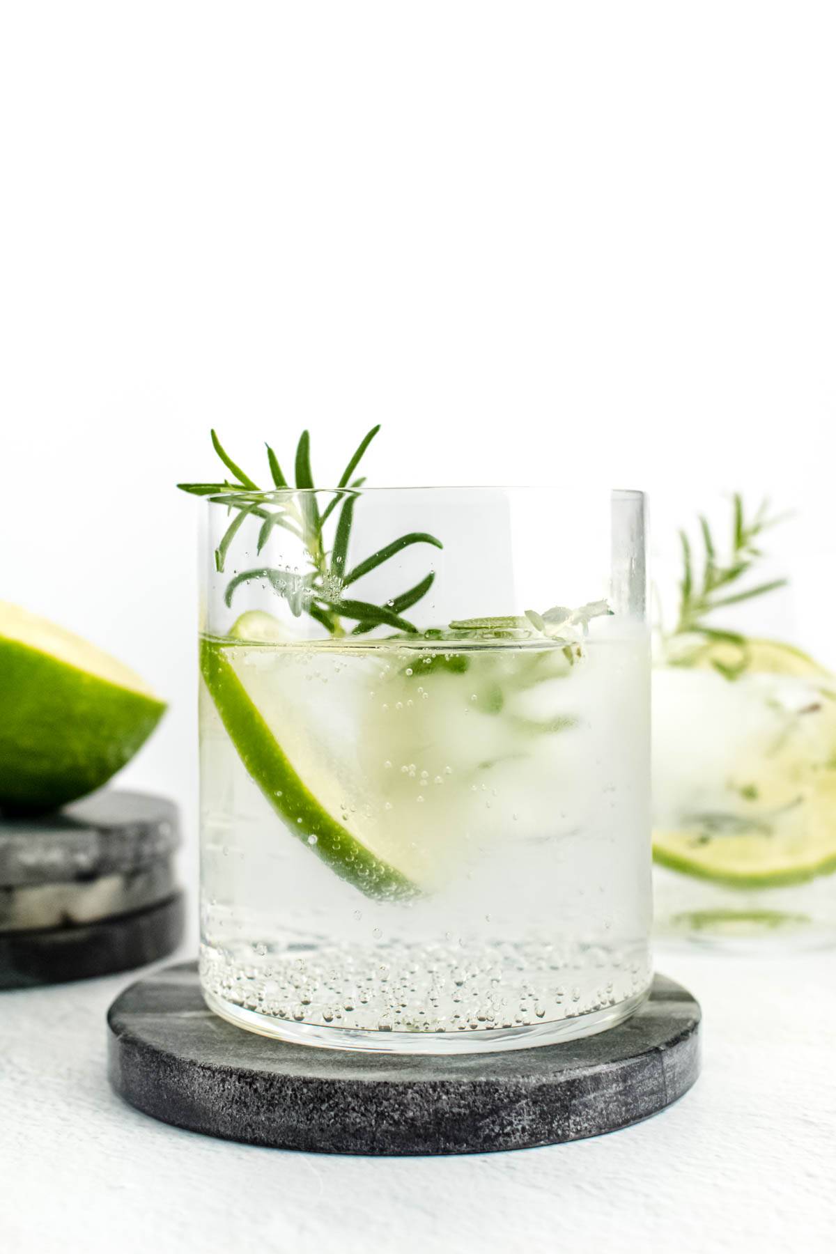 A bubbly clear cocktail sits on a gray stone coaster with a lime round and a rosemary sprig.
