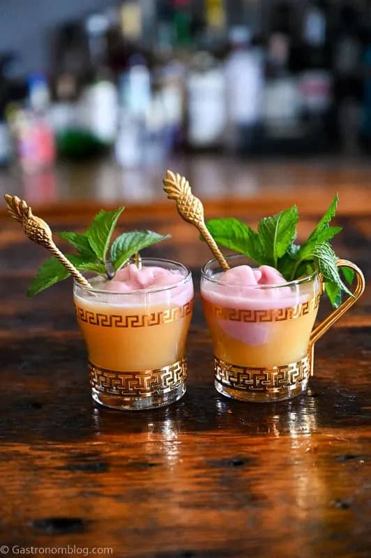 Two glass mugs with gold handles hold an orange drink with scoops of pink sherbet and topped with a mint sprig.