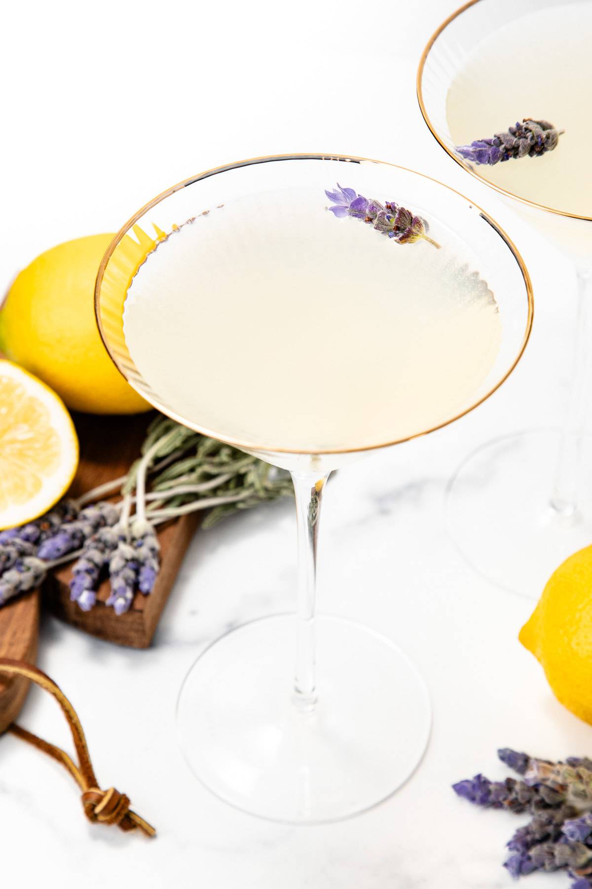 A stemmed glass with a yellow drink garnished with a sprig of lavender near whole lemons and lavender sprigs.