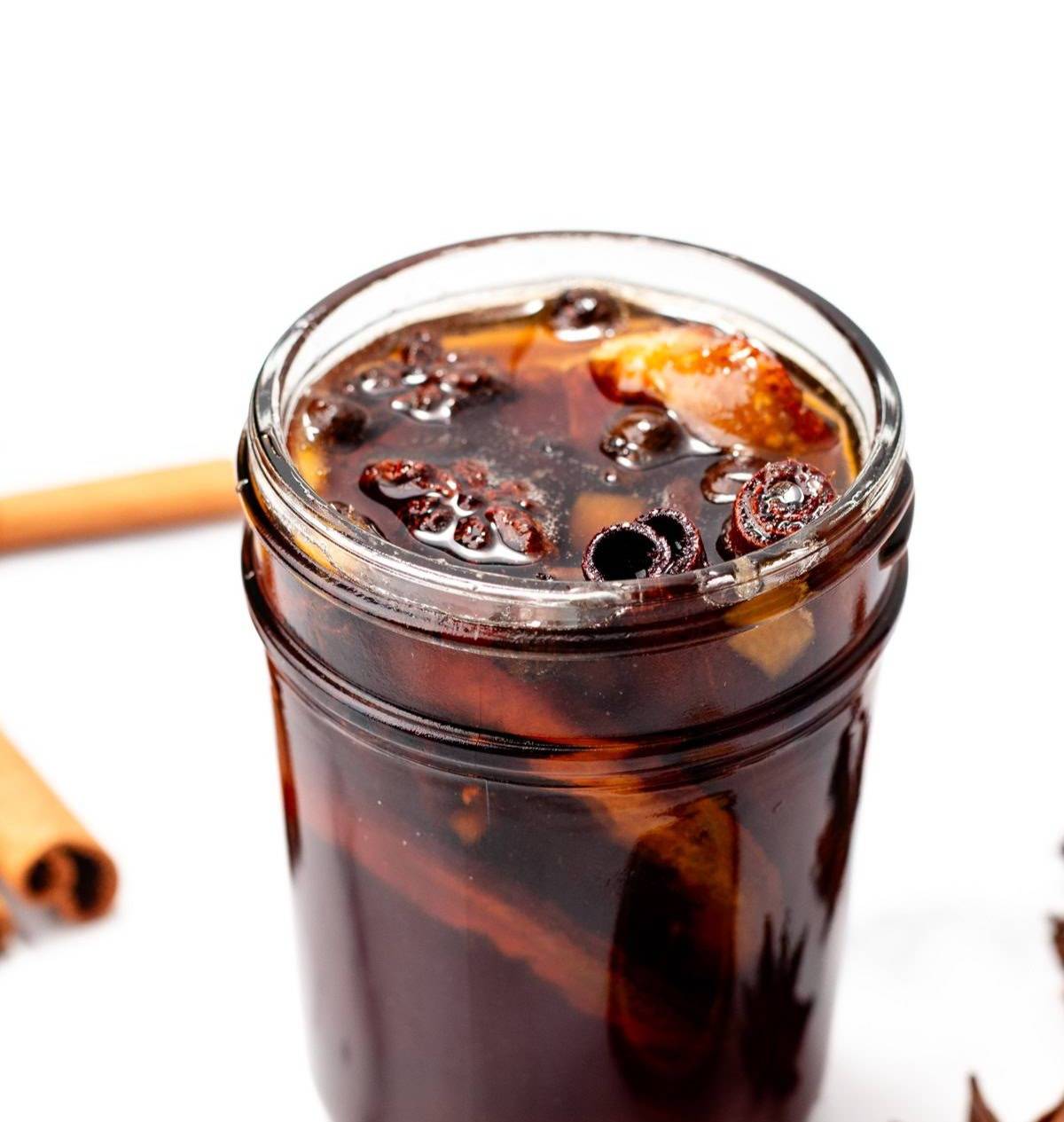A dark brown simple syrup sits in a jar filled with whole spices.