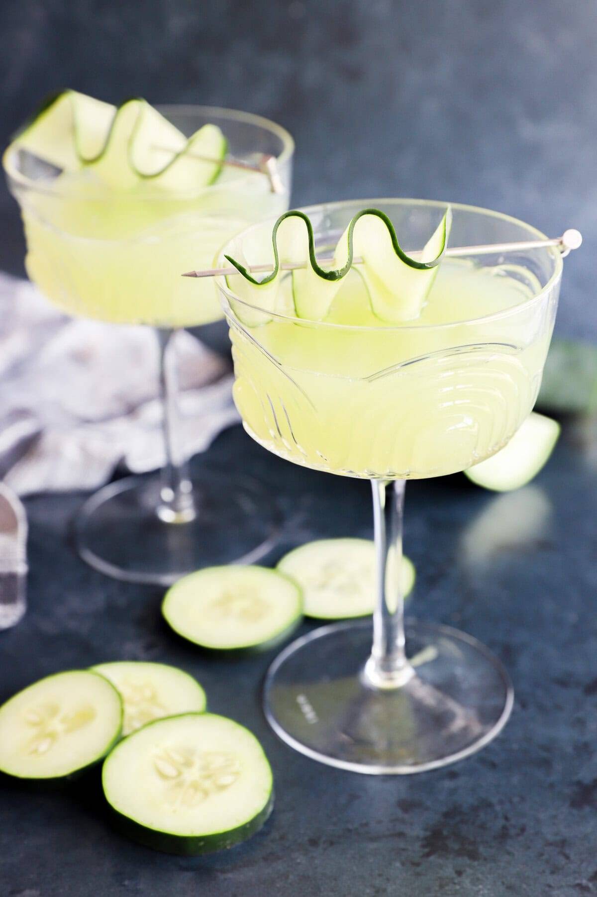 A light green cocktail in a stemmed glass with a cucumber ribbon on a cocktail pick.