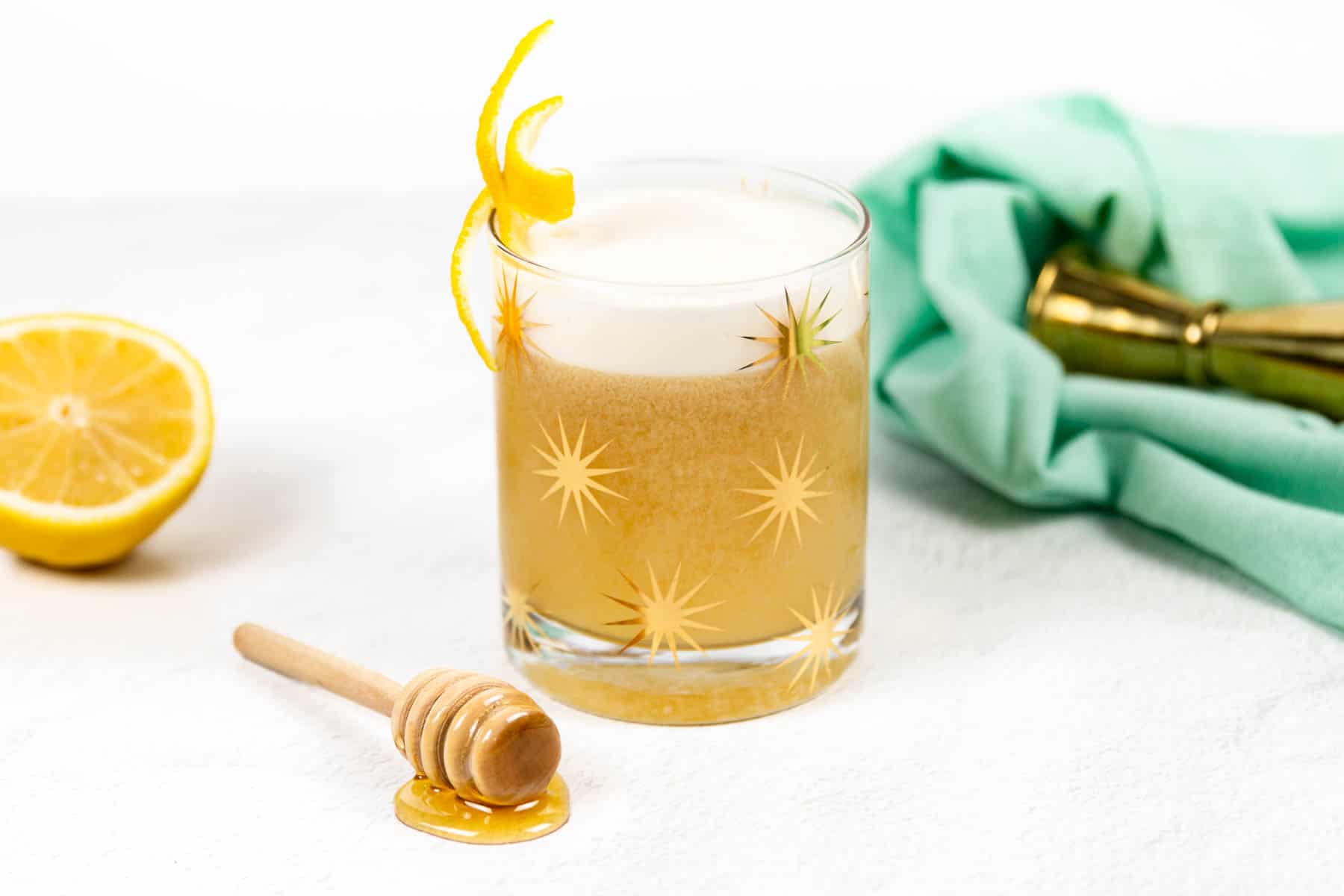 A glass with gold stars holds a pale amber liquid topped with white foam near a honey stirrer.