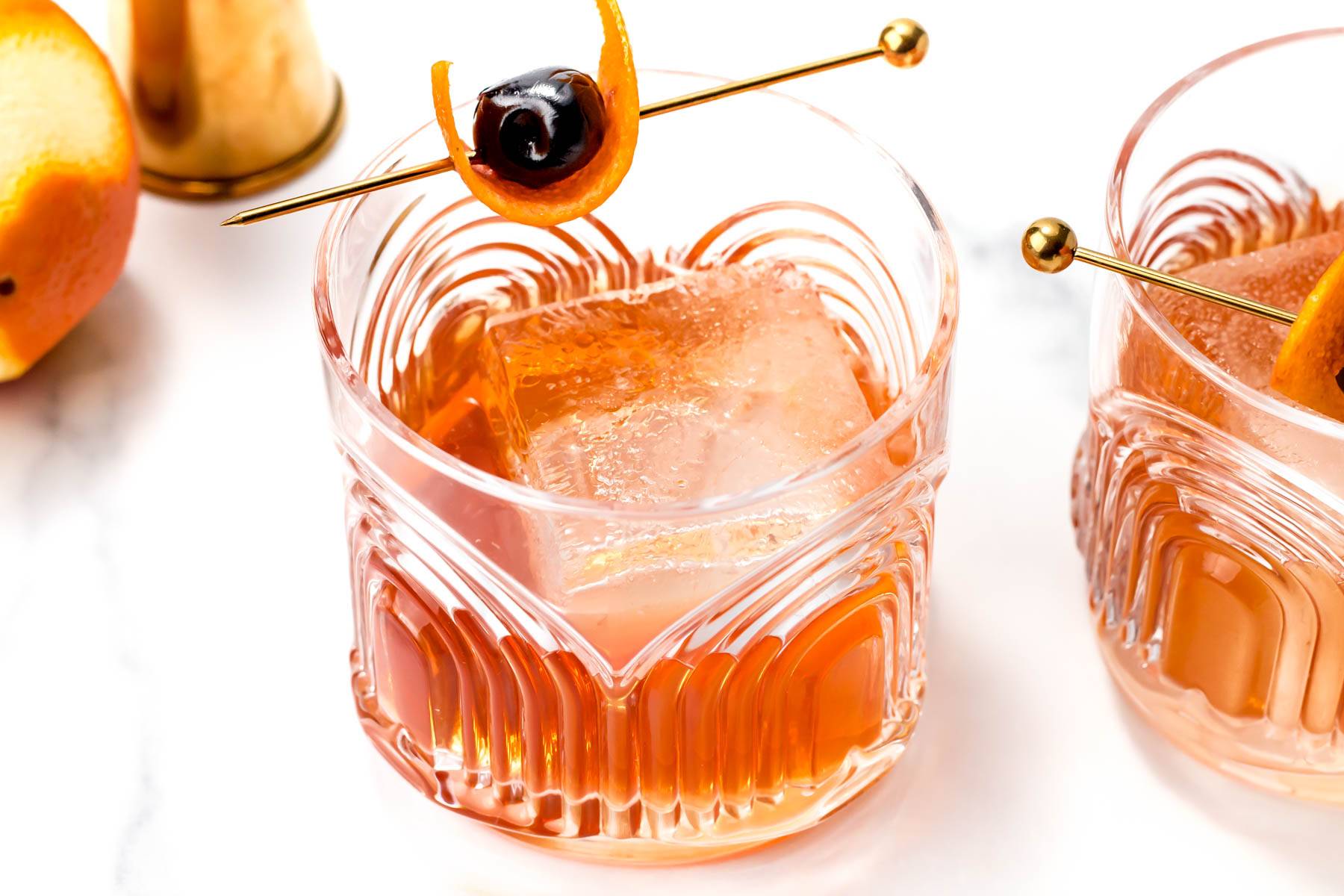 A rocks glass holds an amber liquid with a large square ice cube garnished with a piece of orange peel and cocktail cherry on a gold cocktail skewer.