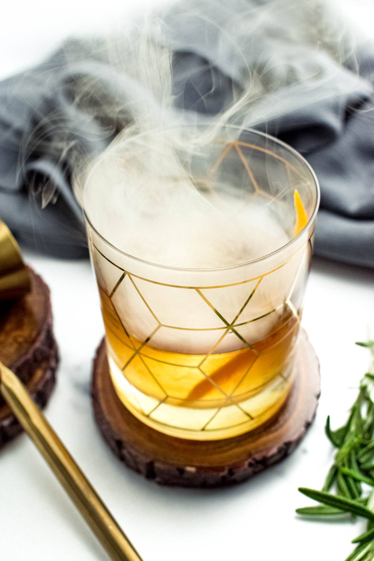 An amber drink in a rocks glass decorated with gold geometric patterns topped with smoke.