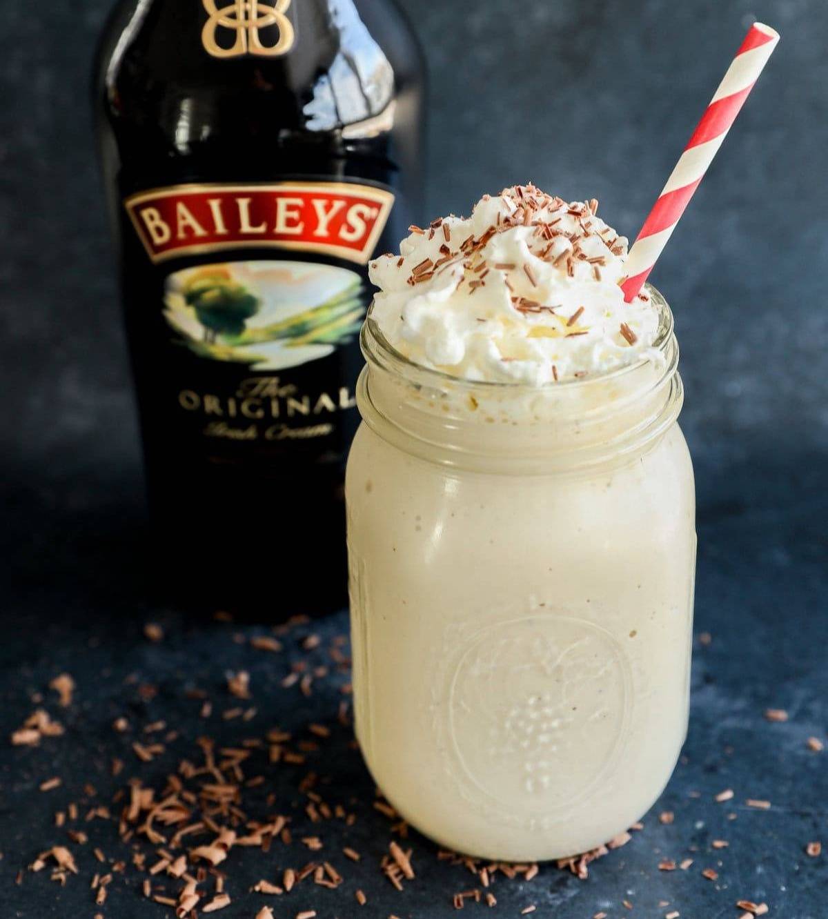 A mason jar holds a pale milkshake topped with whipped cream and chocolate sprinkles in front of a bottle of Baileys.