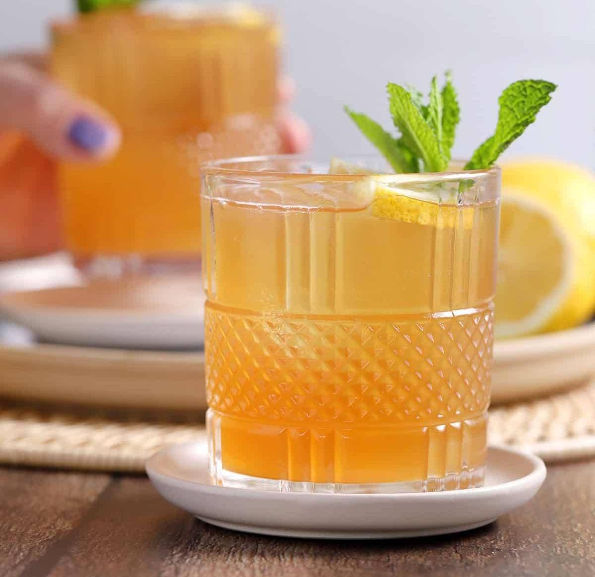 A lemonade and tea cocktail sit in a short glass on a white saucer with a mint sprig garnish.