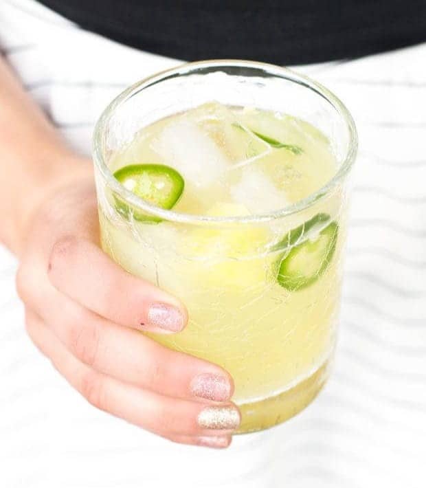 A hand holds a yellow drink with jalapeño slices.