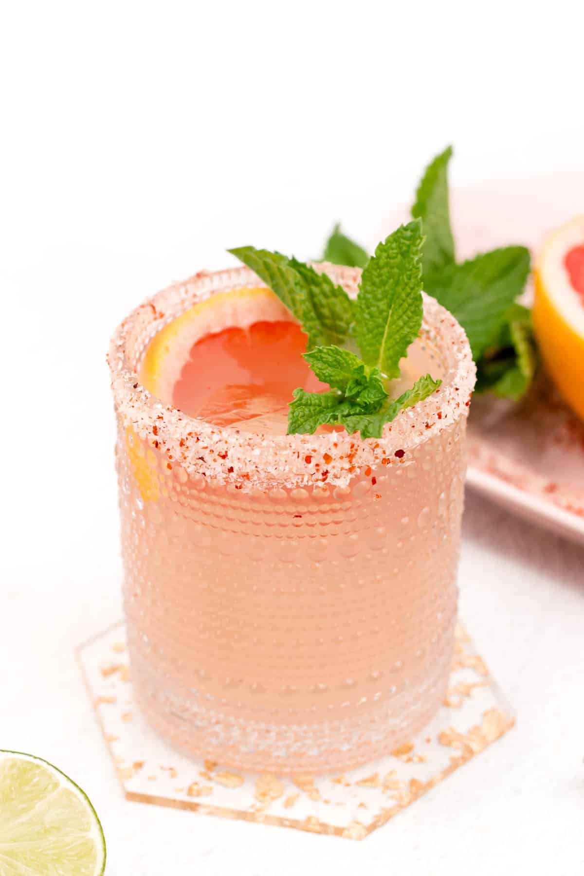 A pink grapefruit cocktail with sugar on the rim garnished with mint leaves and a grapefruit slice.