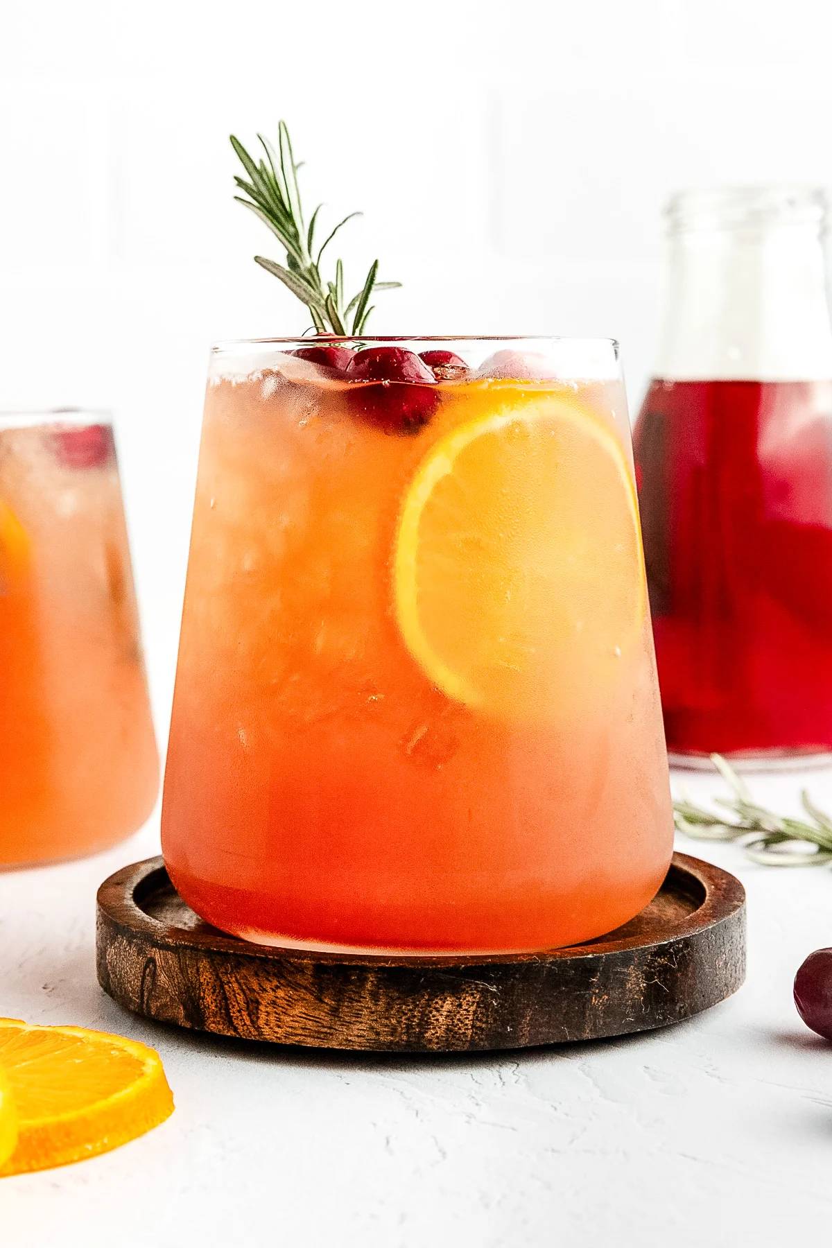 A red-orange drink sits on a round wooden coaster with a slice of orange, whole cranberries and a rosemary sprig on top.