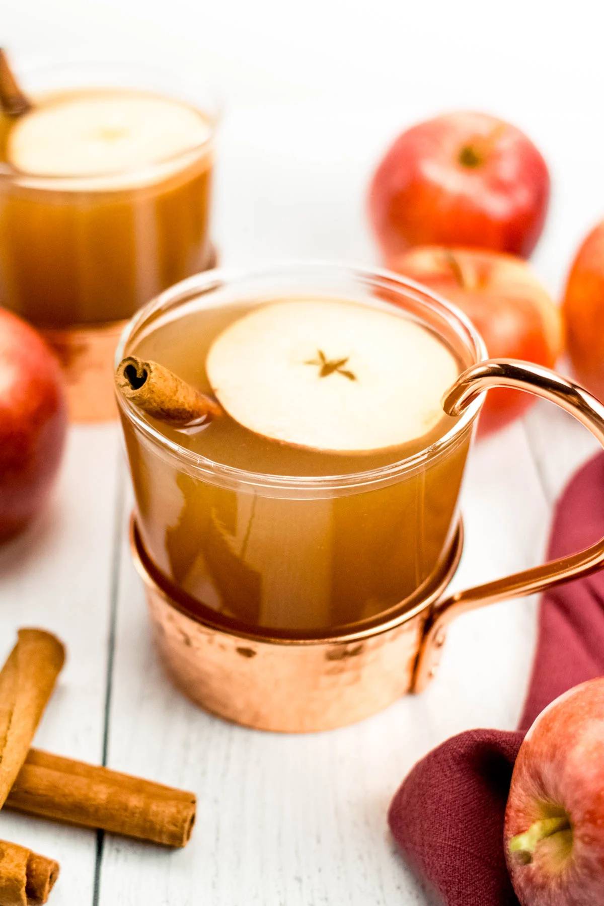 A copper and glass mug holds apple cider topped with an apple slice and cinnamon stick.