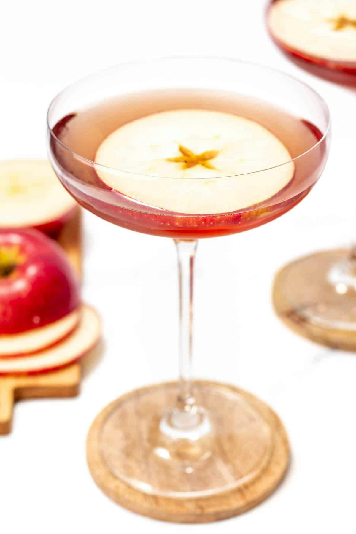 A red cocktail in a coupe glass with an apple round floating on the top.