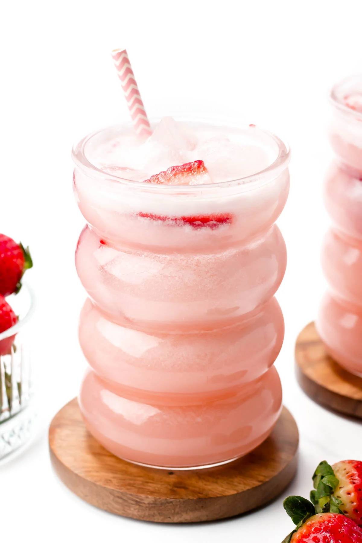 A pink iced drink with floating strawberry slices on a round wooden coaster.