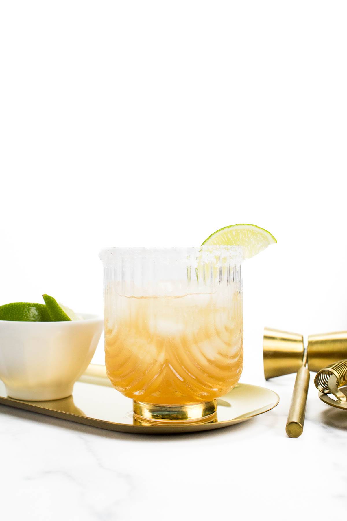 An amber drink in a glass rimmed with salt and a lime wedge sits on an oval gold tray near gold bar tools and a white bowl of lime slices.