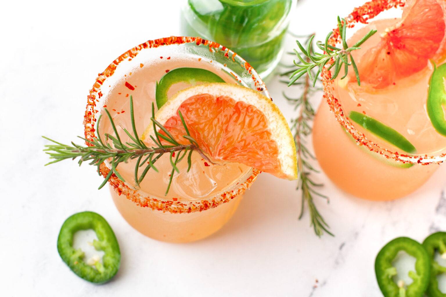 Overhead view of two light pink cocktails with a grapefruit slice, jalapeño slice and rosemary sprig on top.