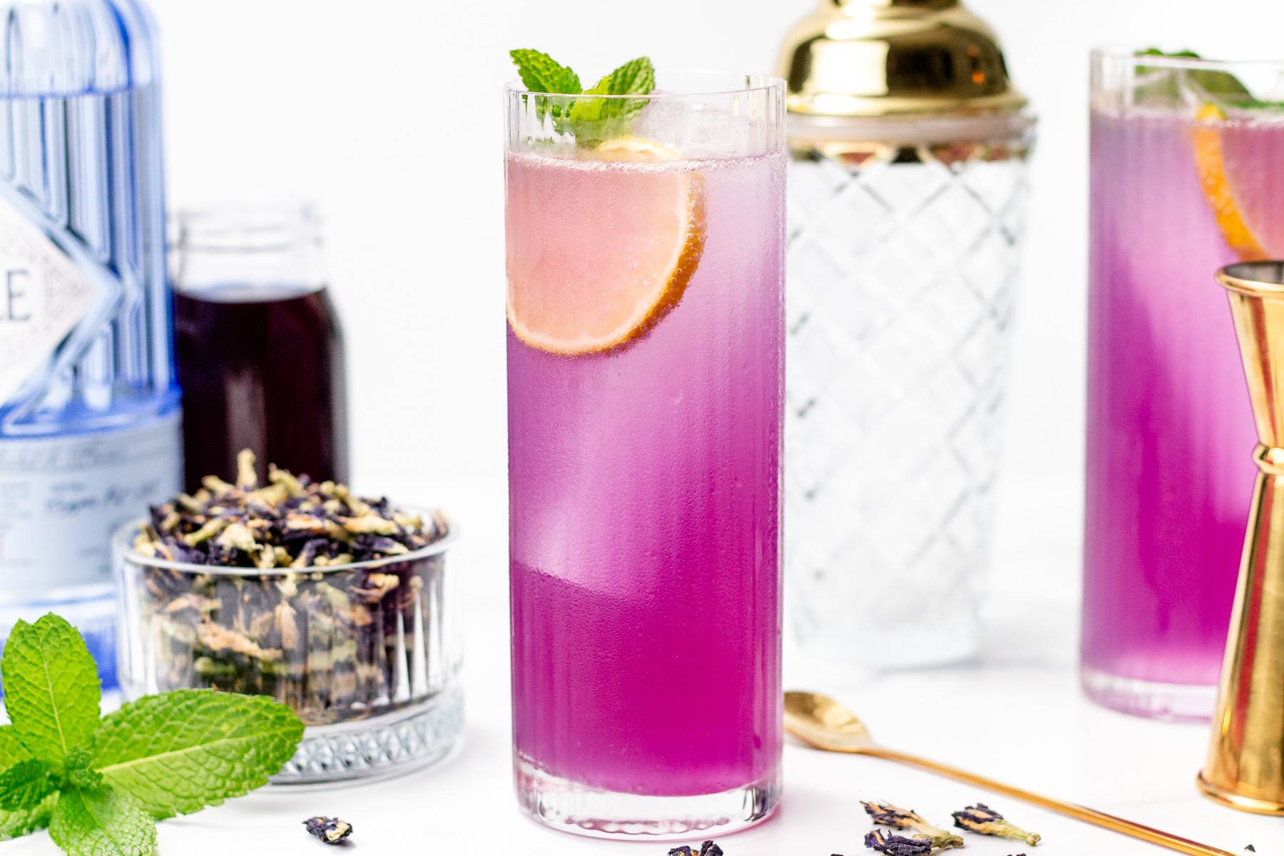 Two purple drinks sit near cocktail tools and a glass bowl of dried purple flowers.