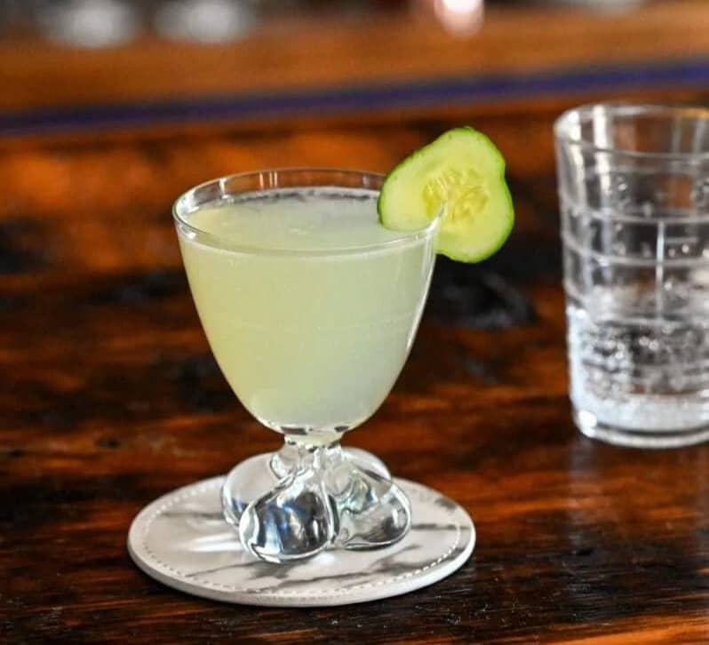 A glass with a pale green drink garnished with a cucumber round.