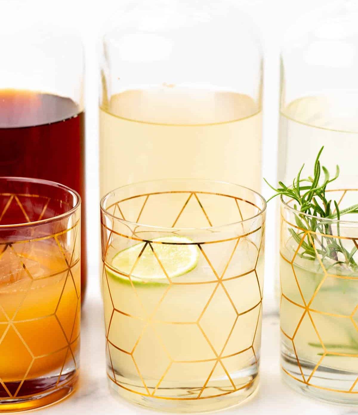 Three glasses holding colorful drinks sit in front of large glass bottles.