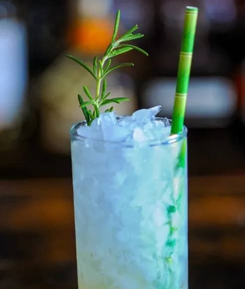 A drink filled with crushed ice and a rosemary sprig and a green straw.