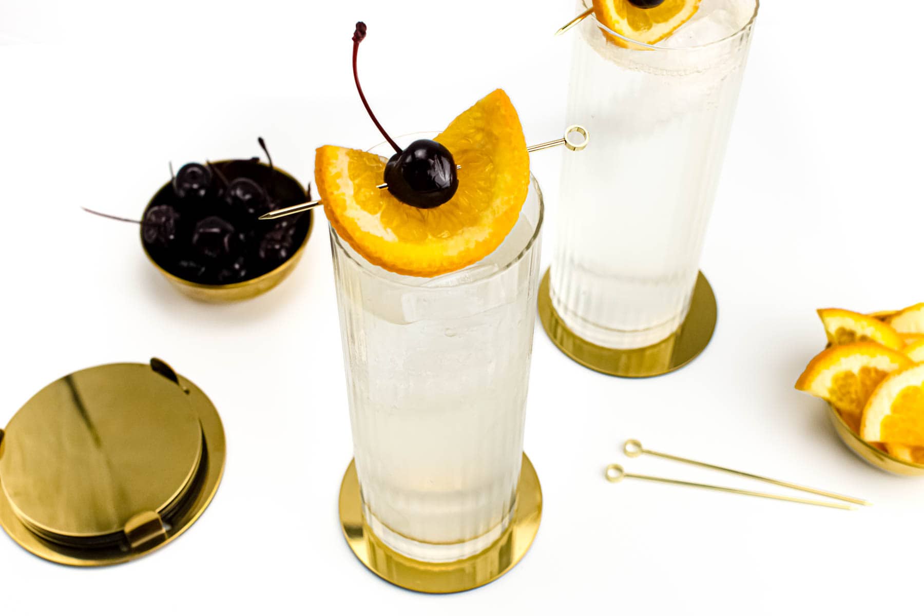 Two clear cocktails in tall glasses with orange slices and cherries as a garnish.