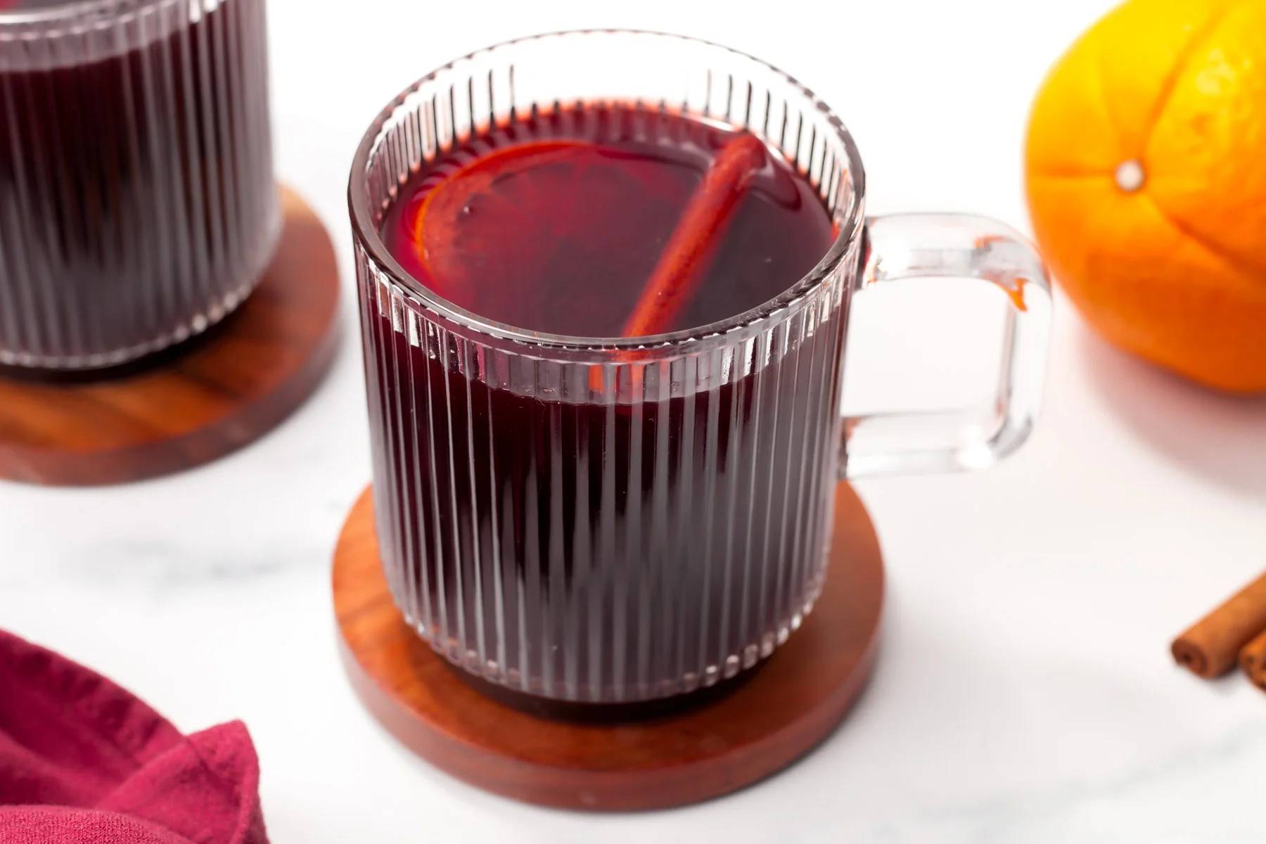 A glass mug holds a deep red liquid garnished with a cinnamon stick and orange round.