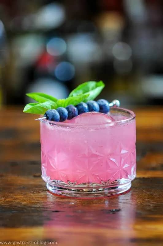 A pink cocktail with a cocktail skewer of blueberries across the top.