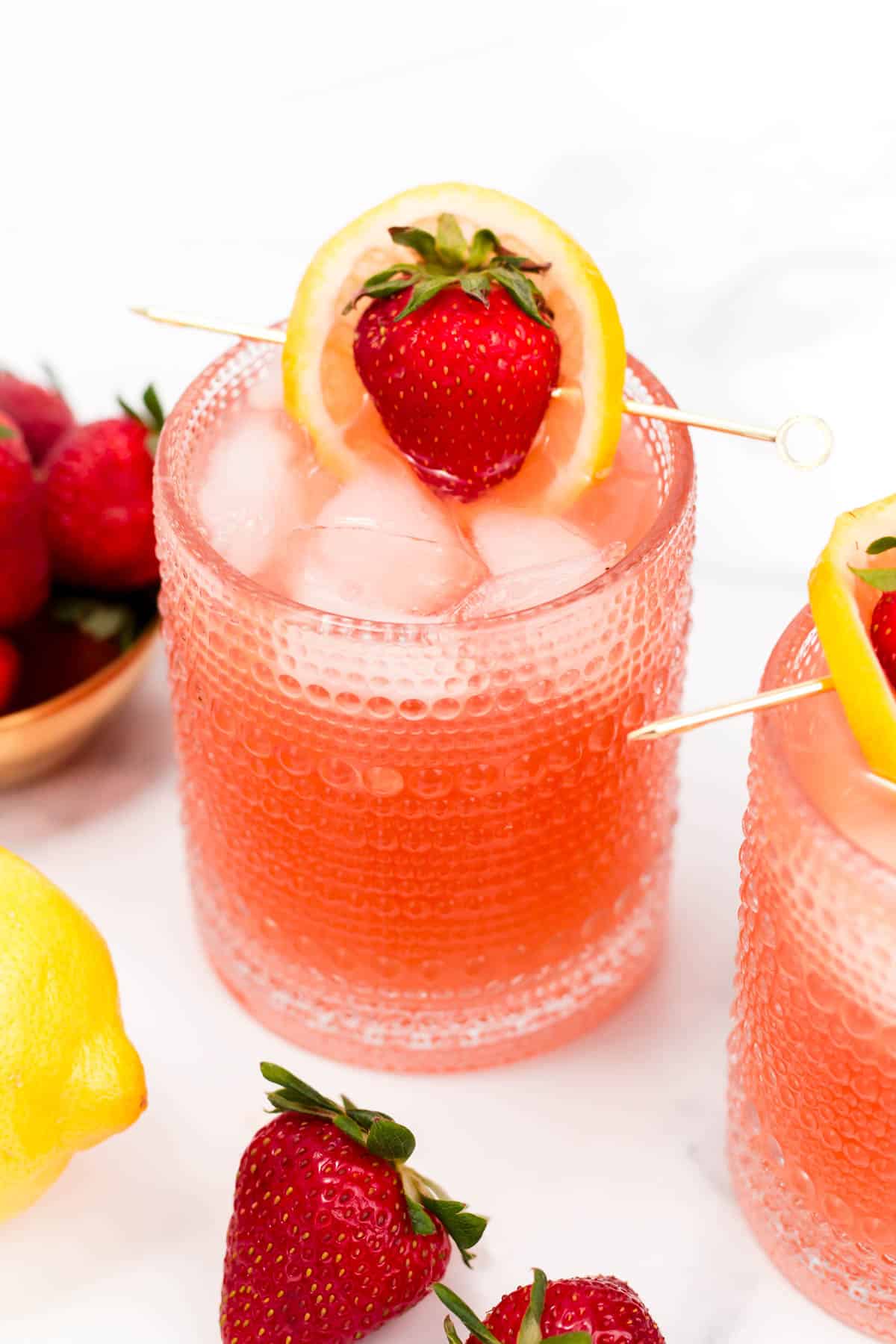 A pink drink topped with a cocktail pick holding a lemon round wrapped around a strawberry.