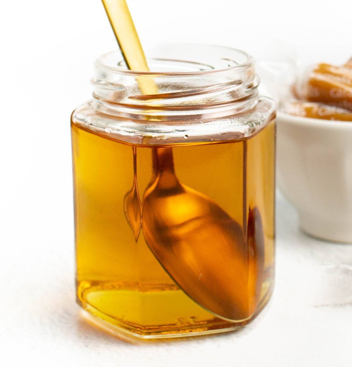A glass jar holds an amber syrup with a gold spoon sitting inside.