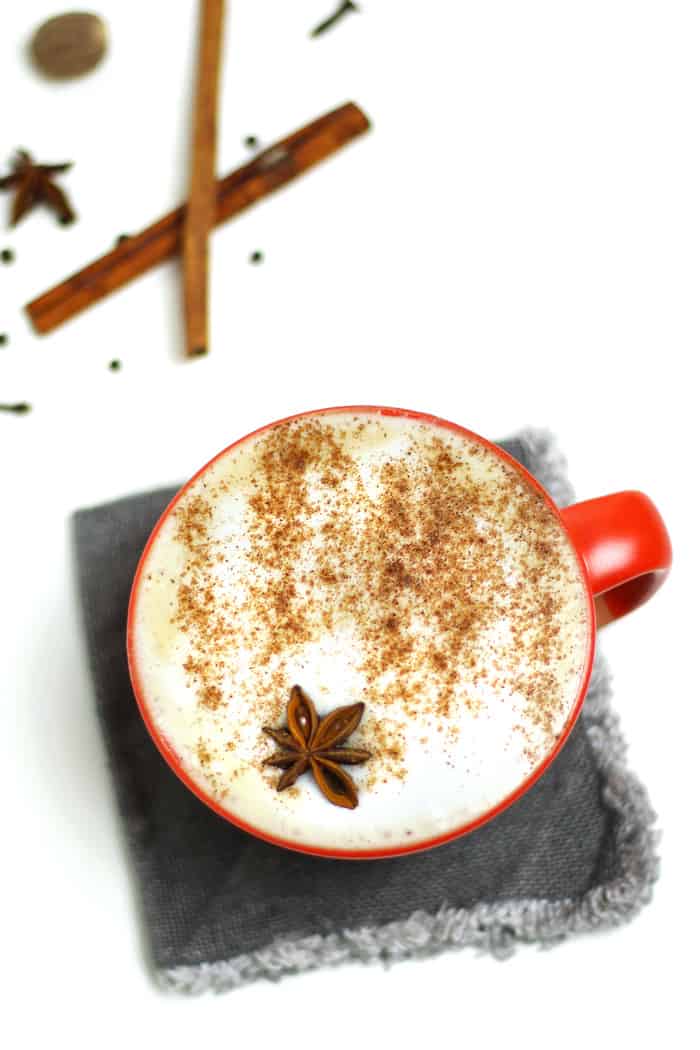 Overhead view of a foamy latte in a red mug topped with a dusting of cinnamon and whole star anise. The mug sits on a charcoal gray square fabric coaster.