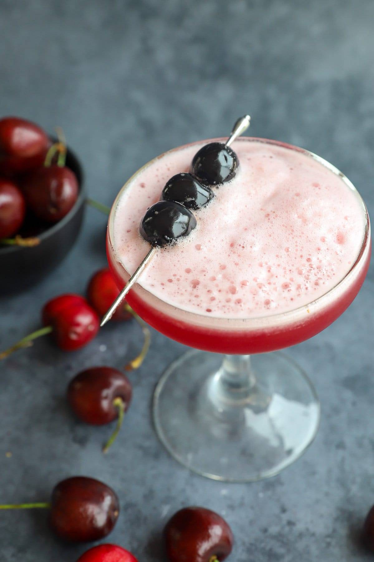 Overhead view of a red foamy drink with a gold pick holding three cherries over the top.