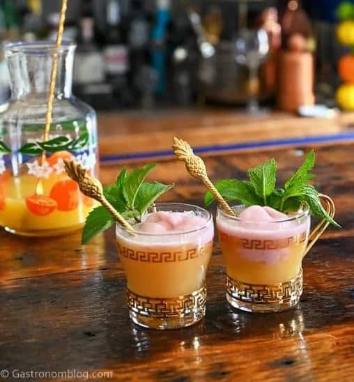 Two glass mugs holding an orange drink topped with pink sherbert with herb sprigs and pineapple skewers.