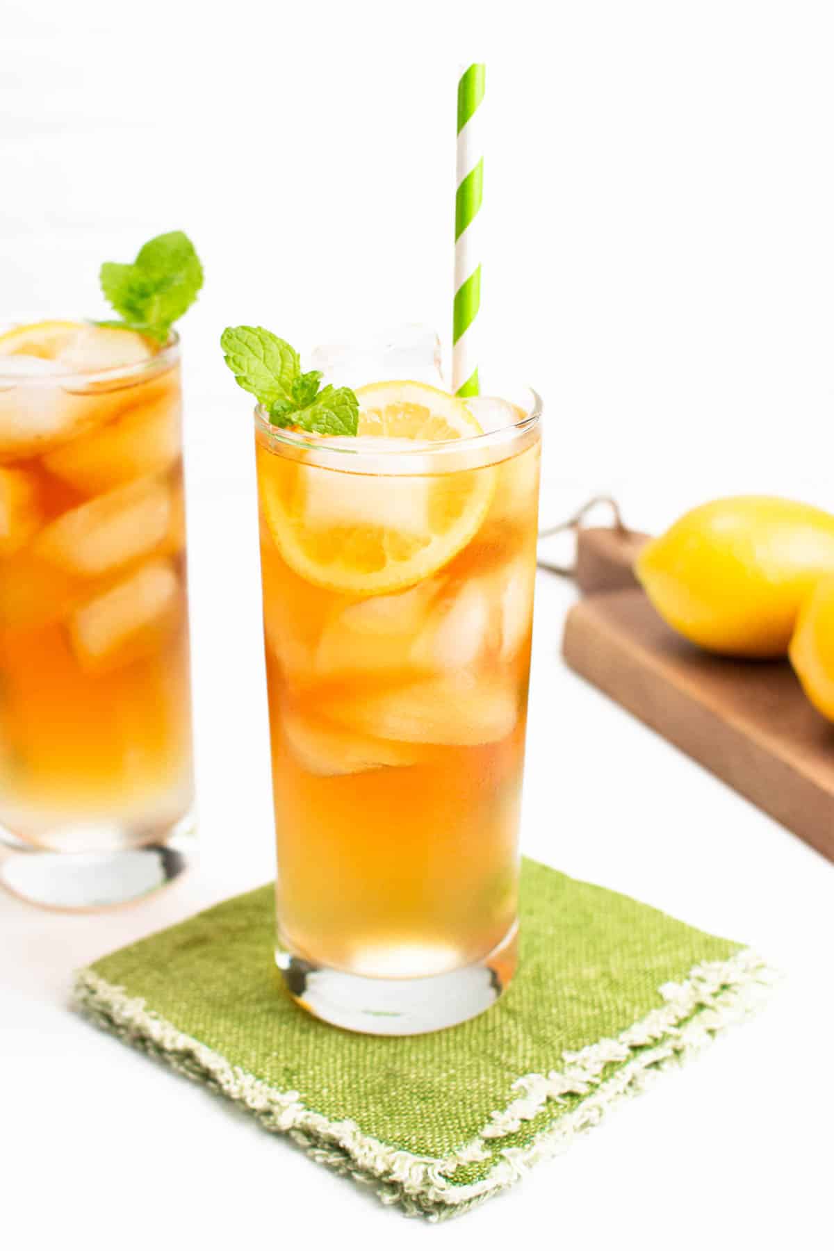 A pale amber drink in a tall clear glass with ice cubes and a lemon wheel sits on a green fabric coaster with a green and white striped straw.