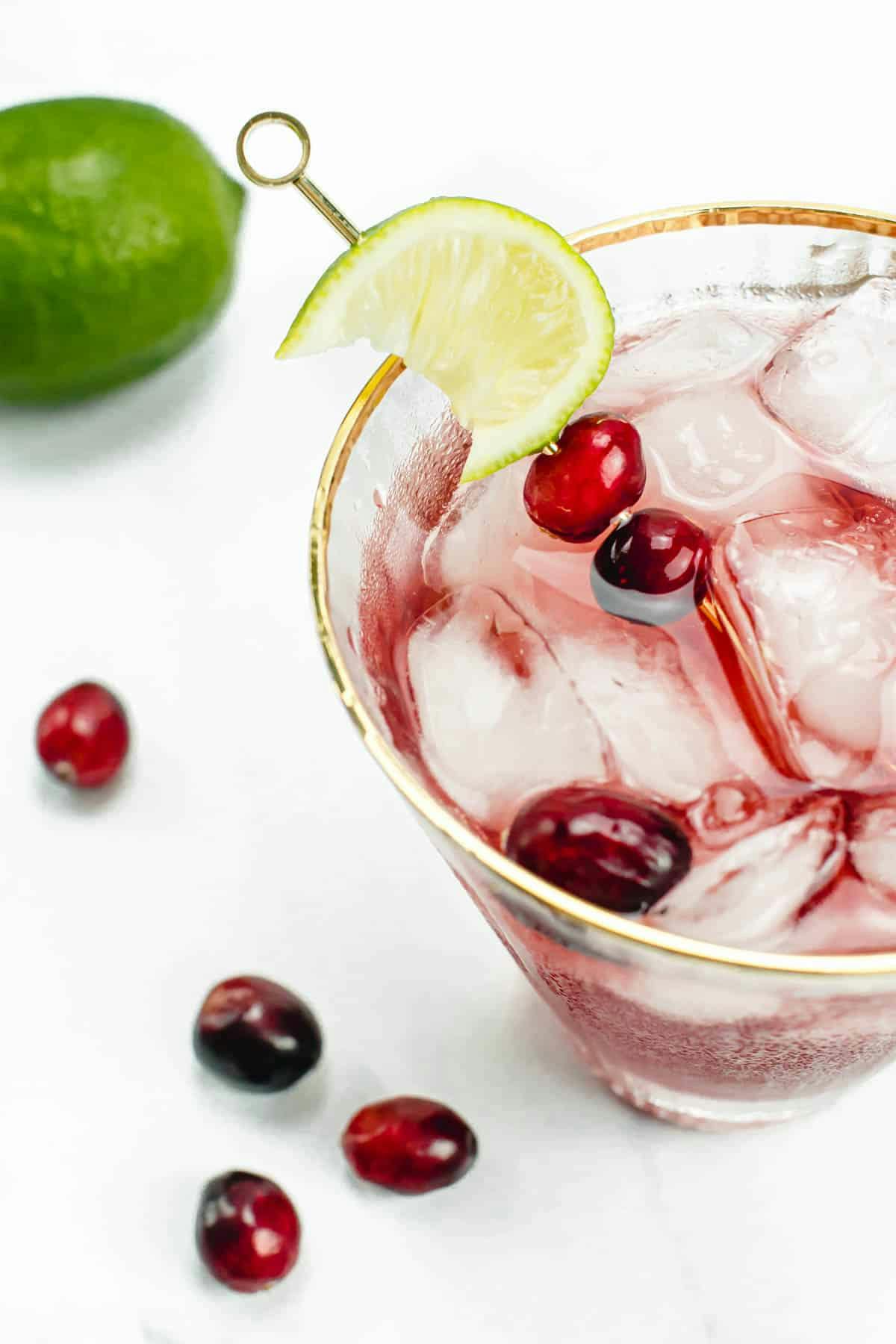 A red cocktail garnished with a lime wedge and whole cranberries on a white surface with several whole cranberries and a whole lime.