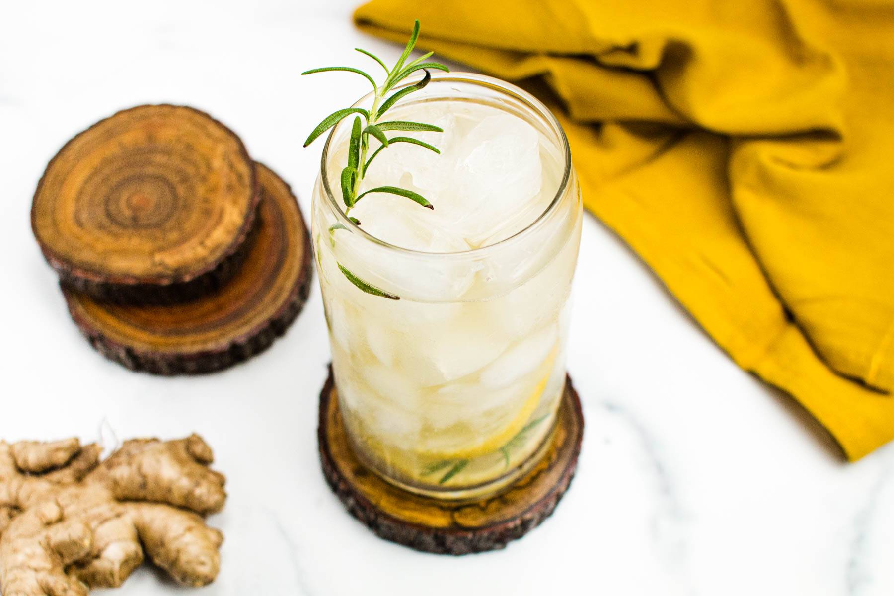 A can glass filled with pale yellow liquid topped with a sprig of rosemary on a round wooden coaster near a ginger root and a mustard colored napkin.