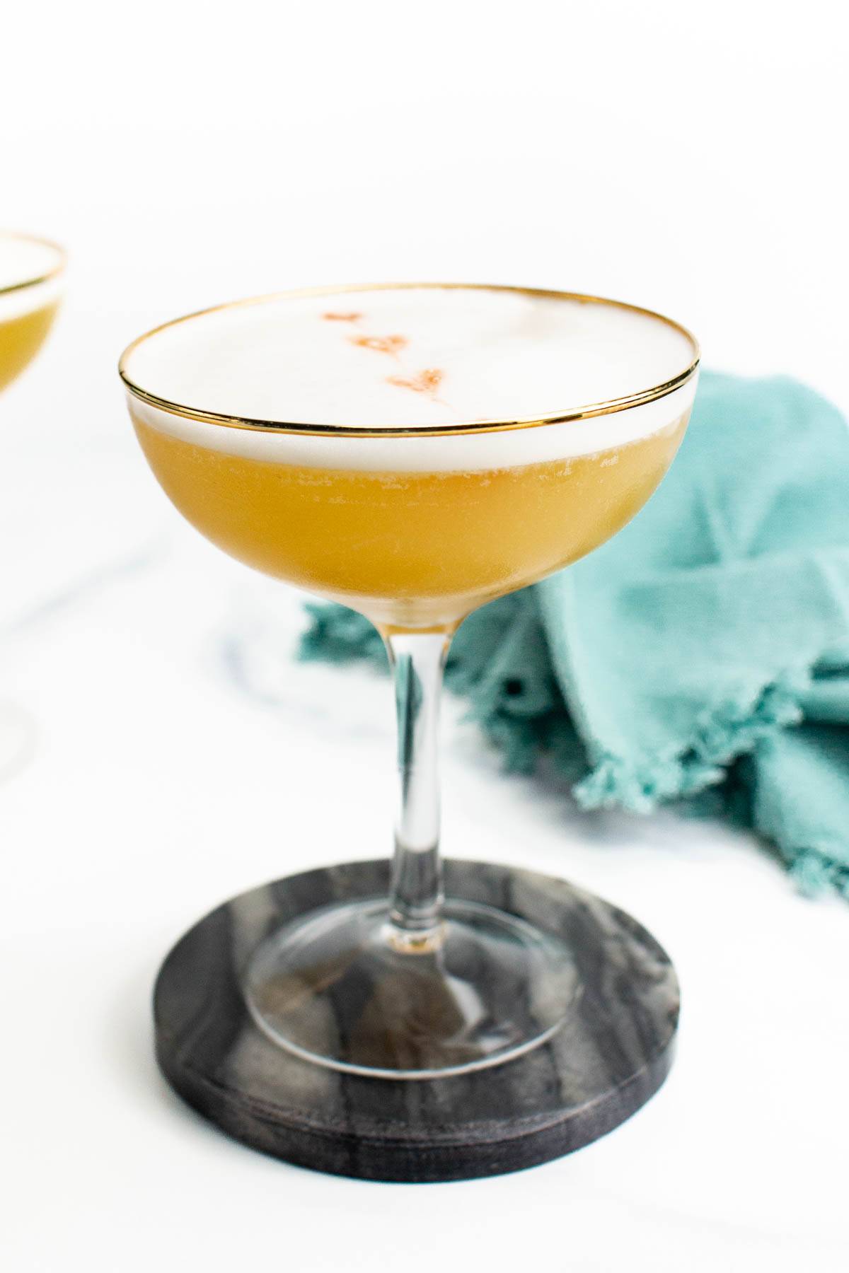 A stemmed glass with a gold rim holds a gold cocktail with a foamy top.