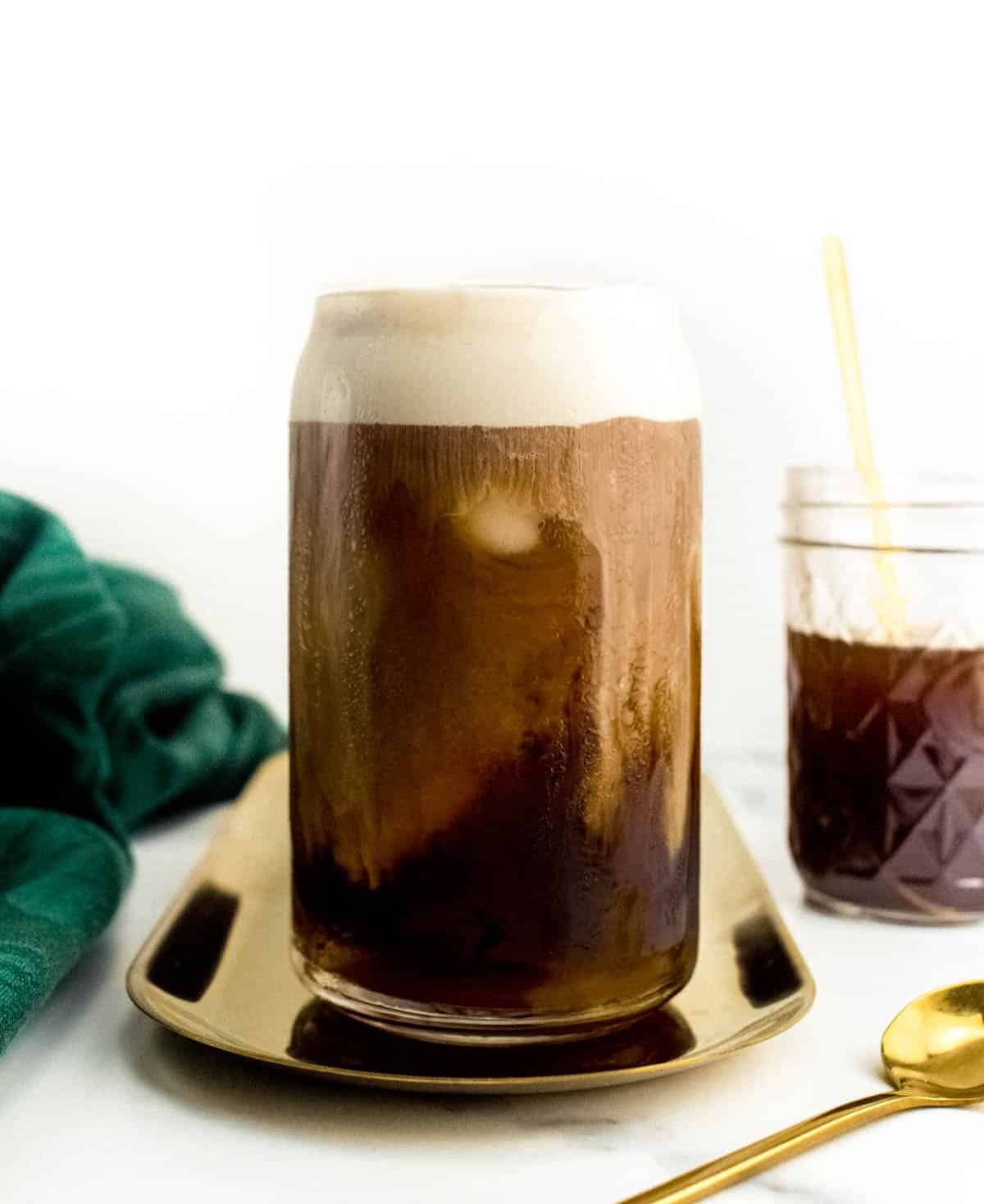 A can glass holds a coffee drink with a foamy top sits on a gold oval tray.
