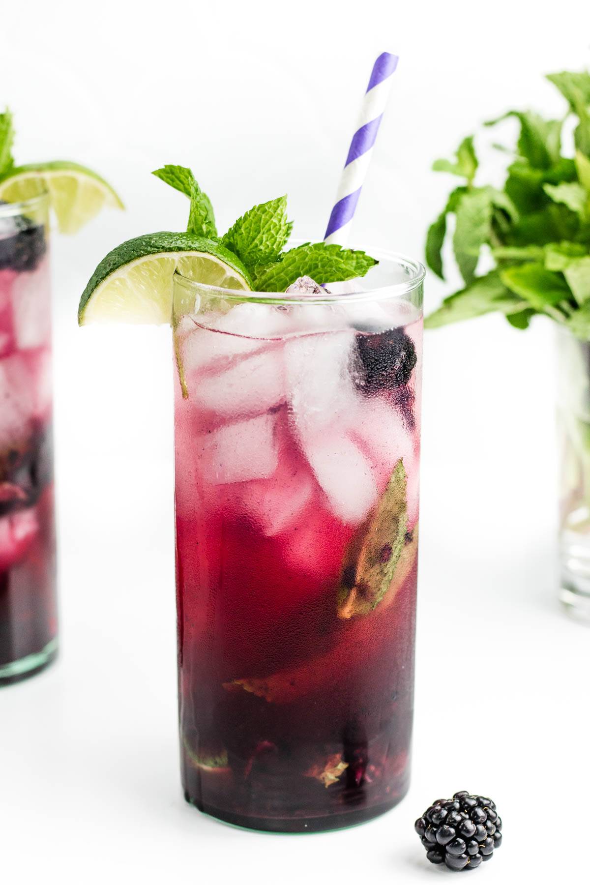 A blackberry cocktail garnished with a lime wedge and mint leaves holds a purple and white striped straw.