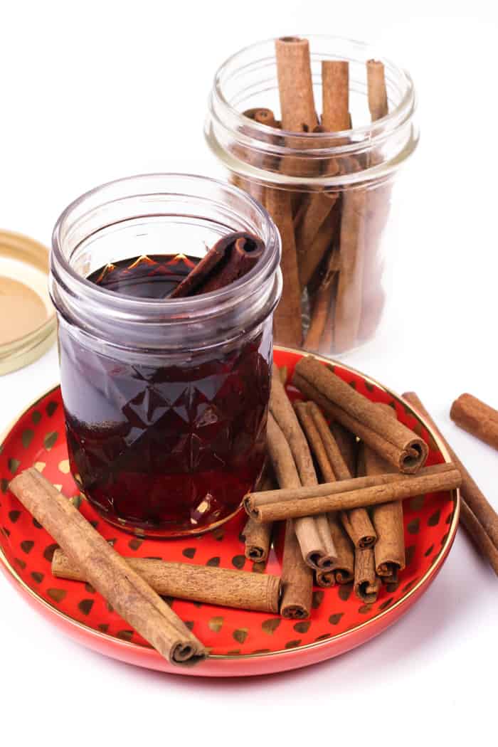 A jar of dark brown liquid with a cinnamon stick sits on a red and gold plate. A full mason jar of cinnamon sticks sits behind.